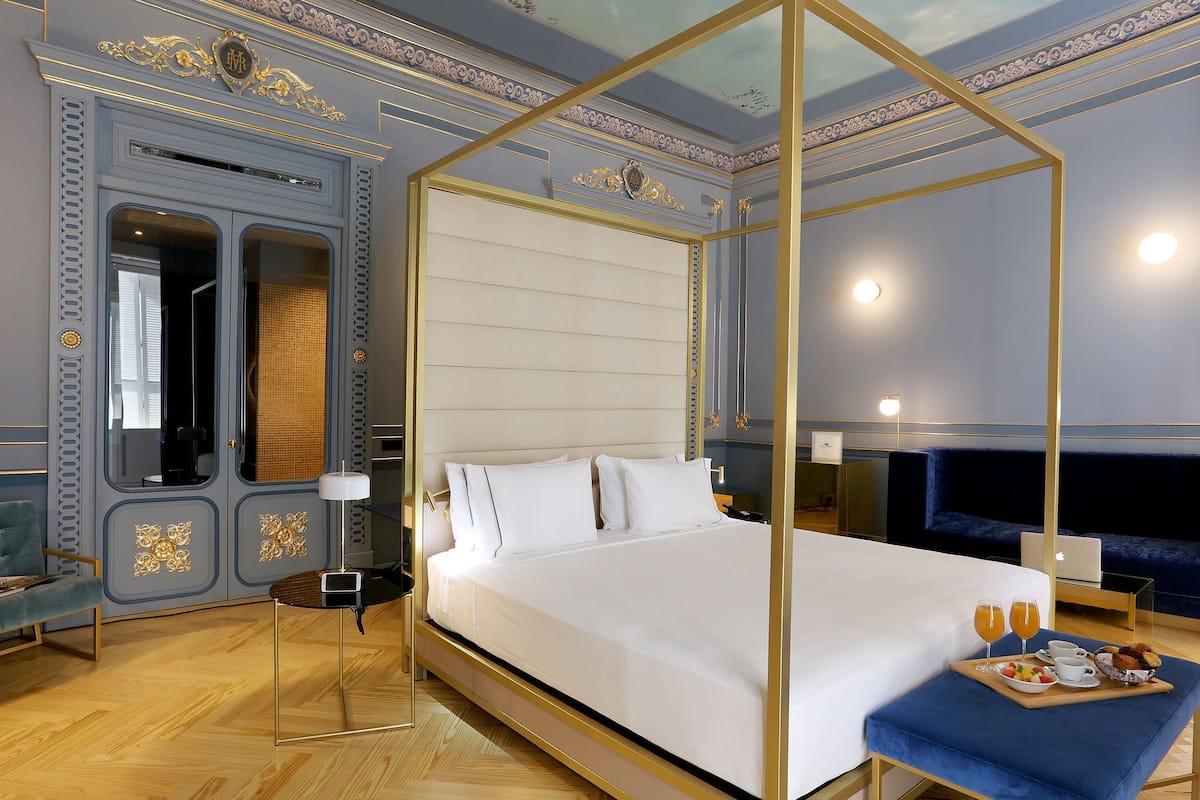 A hotel room with a large bed, a seating area with a blue sofa, a small table with a lamp, and a decorative ceiling.