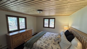 Een slaapkamer met een houten plafond, een bed met beddengoed met een patroon, een dressoir en twee ramen met uitzicht op bomen.