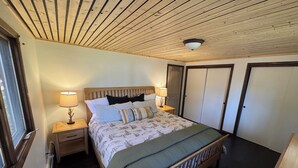 Een slaapkamer met een houten plafond, een bed met beddengoed met een patroon, twee bedlampen, een nachtkastje en een raam.