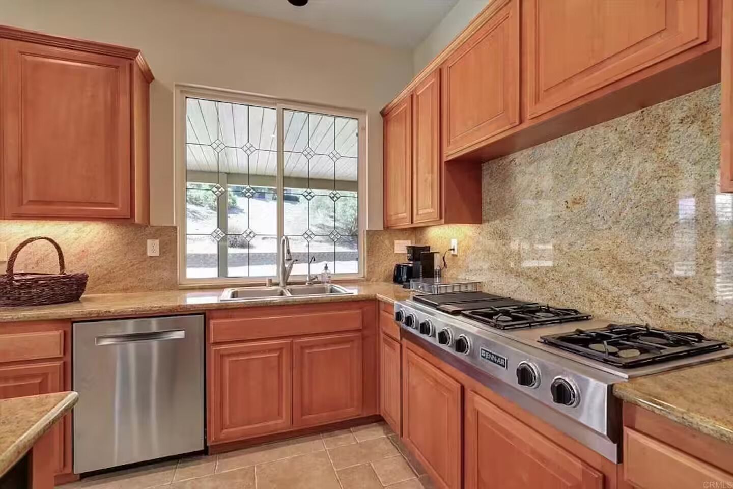 Una cocina con armarios de madera, lavavajillas de acero inoxidable, estufa a gas y ventana con vistas al exterior.