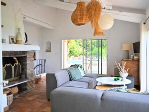 A living room with a fireplace, grey sofa, wicker pendant lights, and a view of greenery outside.