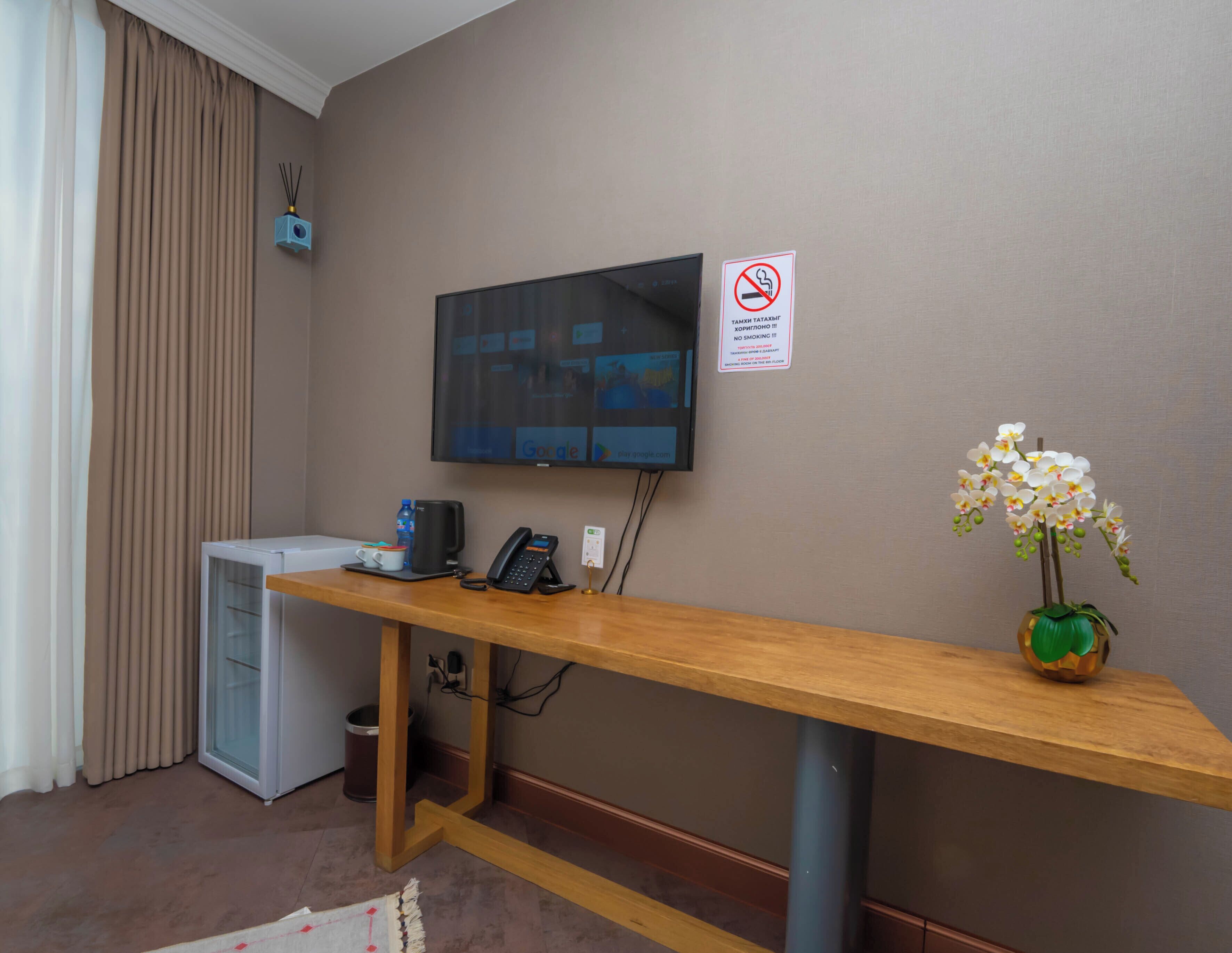 A hotel reception area with a flat-screen TV, a desk with a telephone, a small refrigerator, and a no-smoking sign.