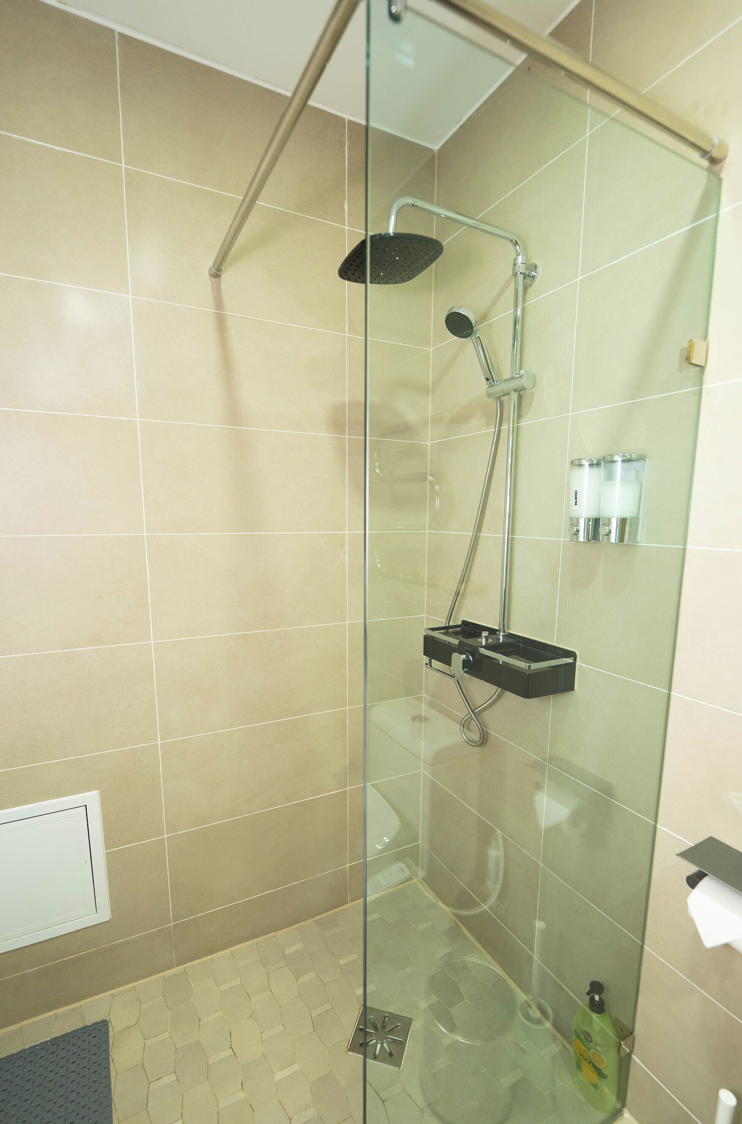 A glass-enclosed shower with a handheld showerhead and a handheld rain showerhead, a glass partition, and a small shelf with toiletries.