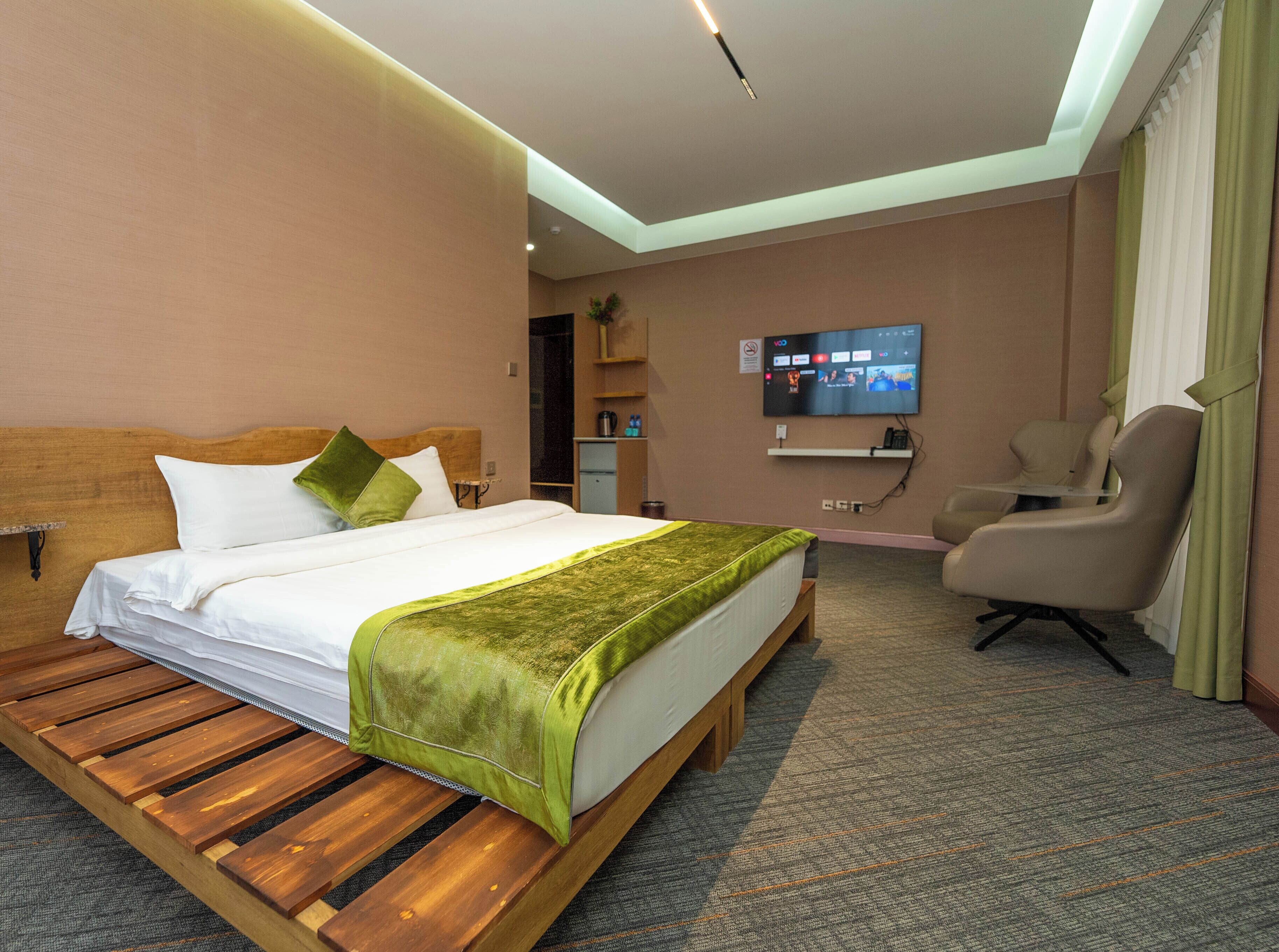 A hotel room with a wooden bed, a green throw blanket, a television on the wall, and a chair.