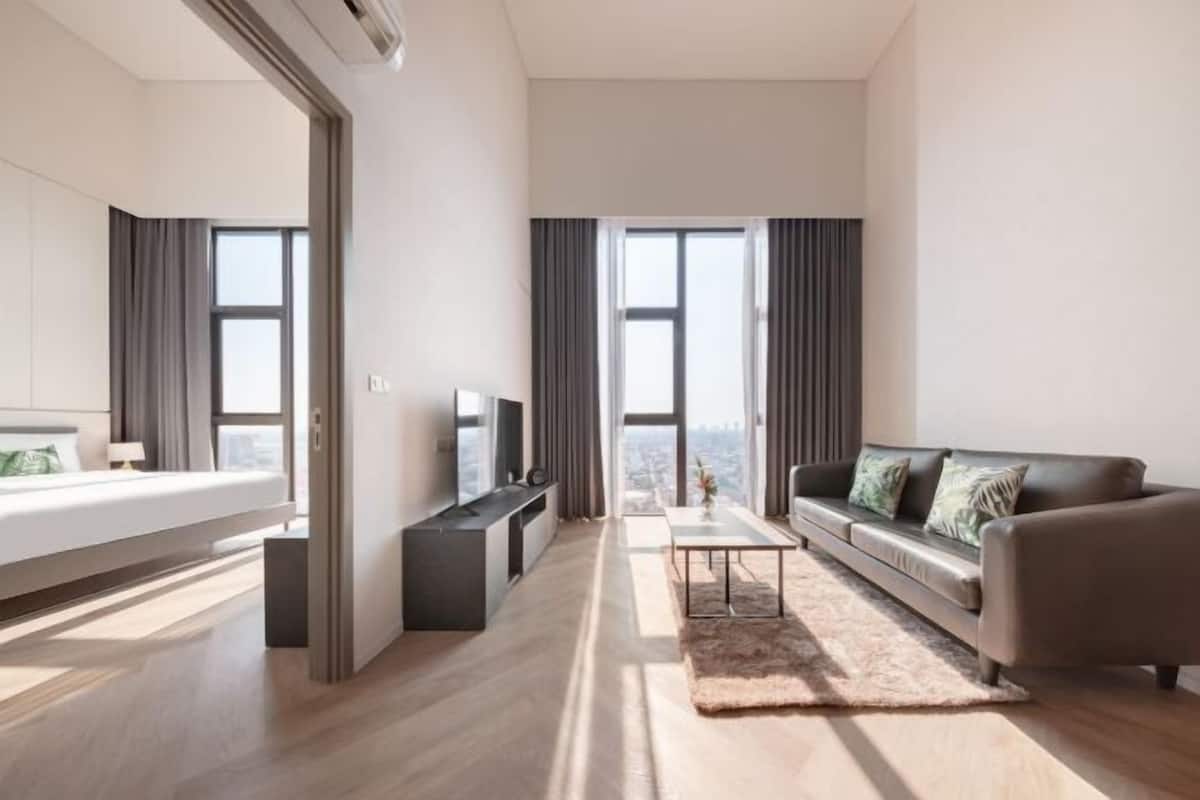 A modern hotel room with a bed, a TV, a sofa, a coffee table, and large windows.