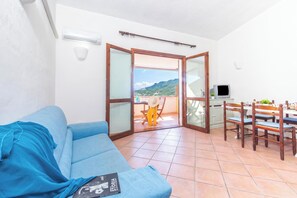 A room with a blue sofa, a dining table with chairs, a television, and a sliding glass door leading to a balcony with a mountain view.