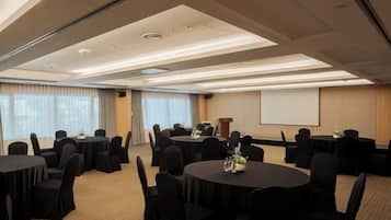 Un salón de conferencias con varias mesas redondas, sillas negras y una pantalla de proyector en la pared.