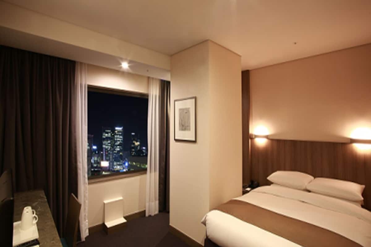 A hotel room with a bed, a desk with a chair, a window with curtains, a framed picture on the wall, and a city view at night.