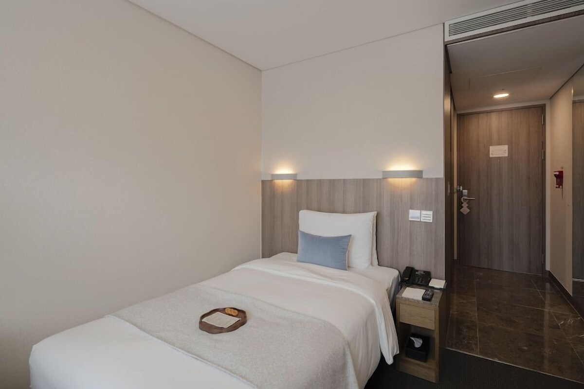 Una habitación de hotel con una cama, mesita de noche y una puerta que da a otra estancia.
