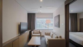 Habitación de hotel con televisor de pantalla plana, sofá, mesa de centro, cama y una ventana grande con vistas a edificios.