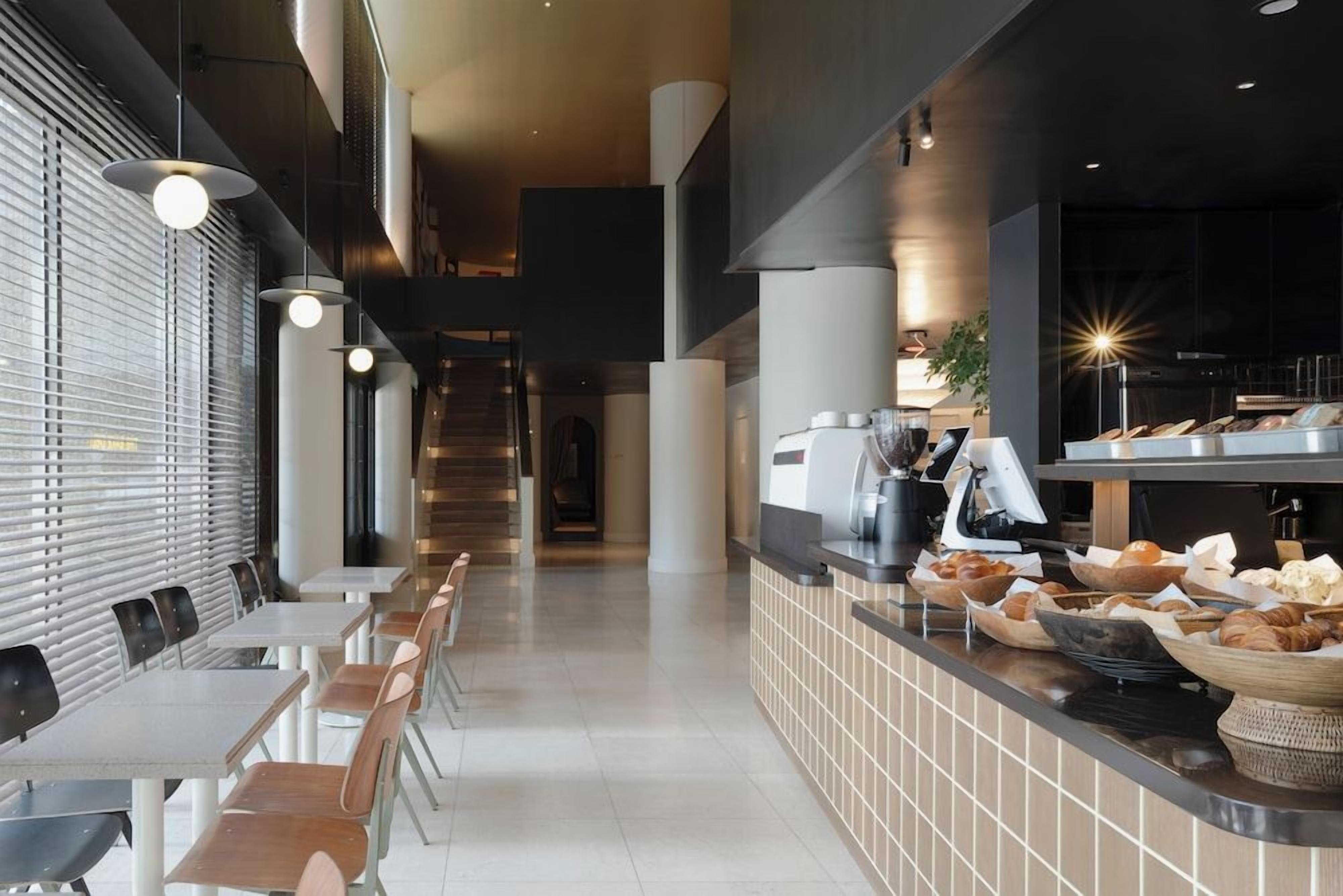 Un café moderno con una barra larga, zona de estar y una escalera al fondo.