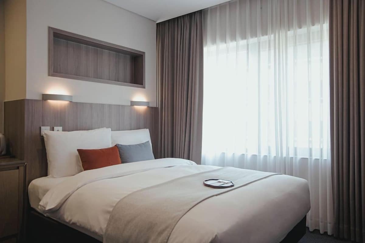 A hotel room with a bed, a nightstand, a built-in shelf above the bed, and a large window with curtains.