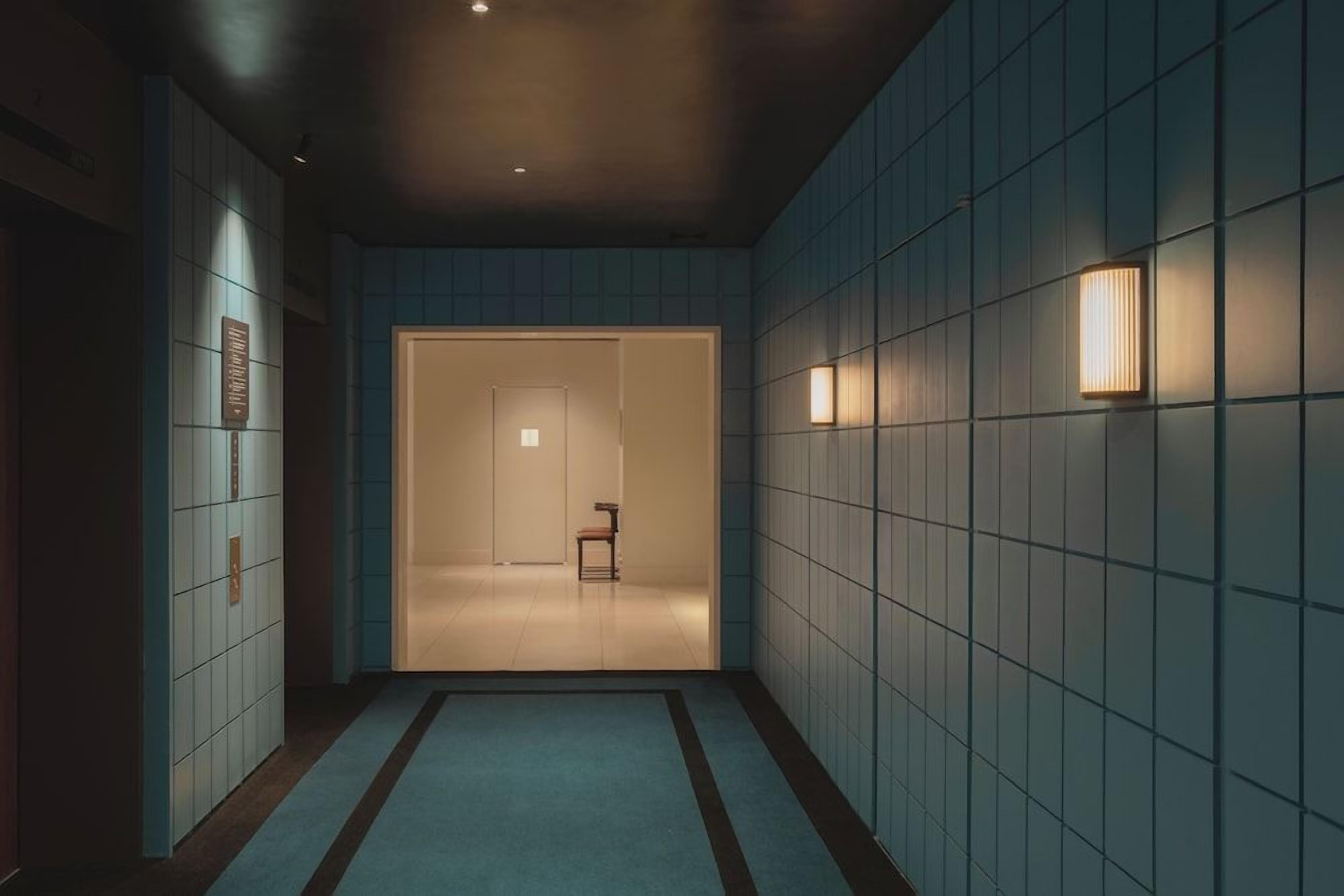 Un pasillo con pared de azulejos, una puerta al fondo y una pequeña mesa con una silla.
