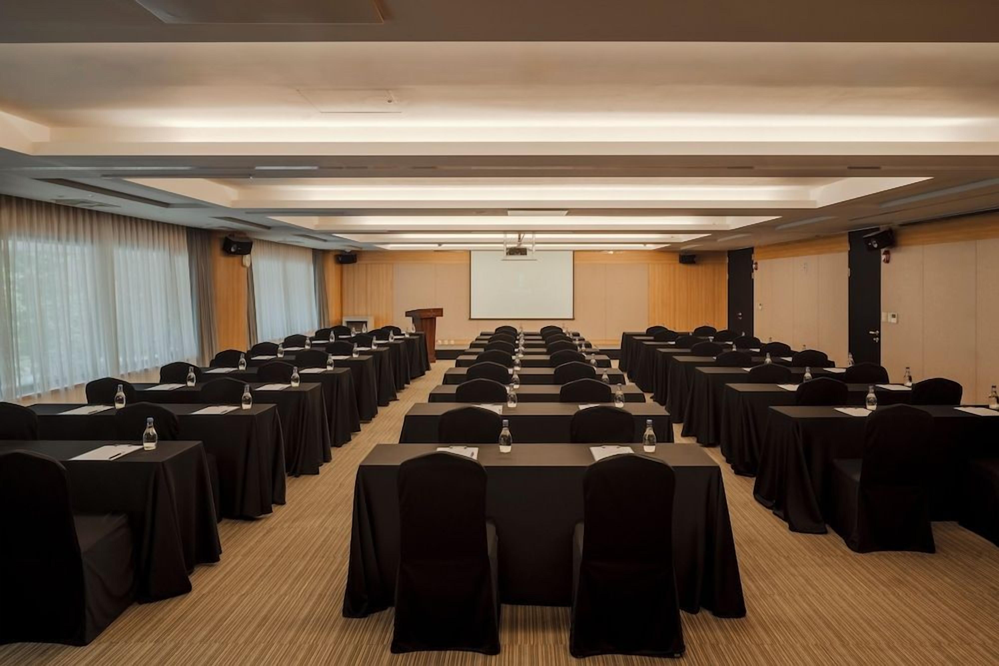 Un salón de conferencias con filas de mesas y sillas, un atril y una pantalla de proyección.