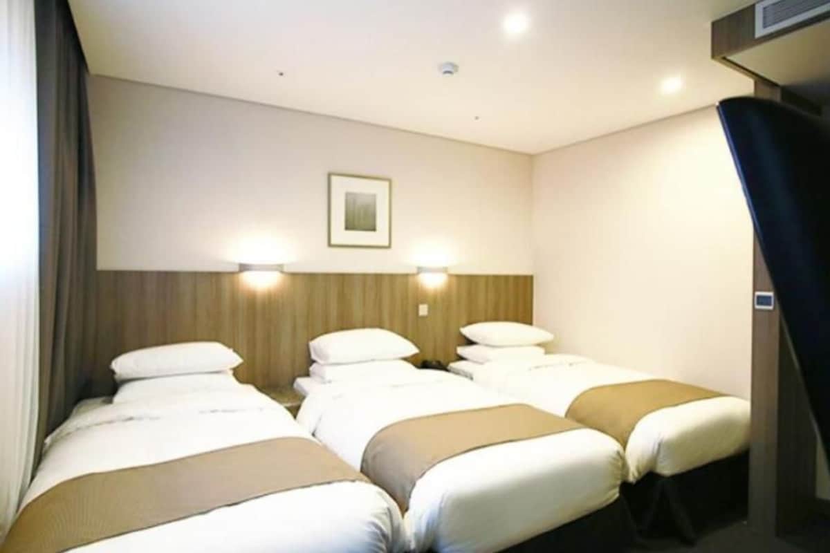 A hotel room with three beds, a wooden headboard, two wall-mounted lights, and a framed picture on the wall.