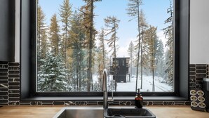 A kitchen with a stainless steel sink, a window with a view of a snowy forest, and a black tiled backsplash. - Off Grid Stays - Treehouse #4 (Columbia Falls)