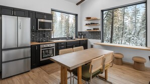 A modern kitchen with black cabinets, stainless steel appliances, a wooden dining table, and a large window with a forest view. - Off Grid Stays - Treehouse #4 (Columbia Falls)