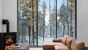 A modern living room with a large window overlooking a snowy forest, a fireplace, and a leather sofa. - Off Grid Stays - Treehouse #4 (Columbia Falls)