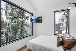 Ein Schlafzimmer mit großen Fenstern, einem Bett mit weißer Bettwäsche, einem braunen Kopfkissen und einem an der Wand befestigten Fernseher.
