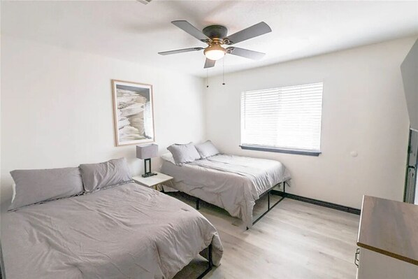 Un dormitorio con dos camas, ventilador de techo, ventana con persianas y un cuadro en la pared.