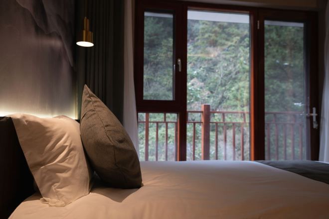 Un dormitorio con una cama grande, dos almohadas, una mesita de noche y una ventana con vistas a árboles.