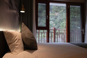 Een slaapkamer met een groot bed, twee kussens, een nachtkastje en een raam met uitzicht op bomen.