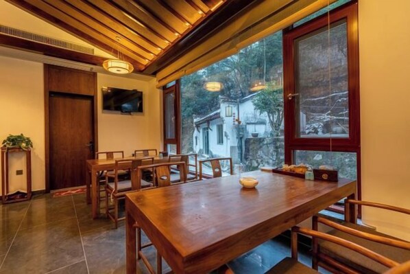 Un coin repas avec une table et des chaises en bois, un téléviseur fixé au mur et de grandes fenêtres donnant sur l’extérieur.