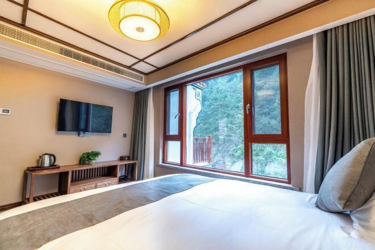 A hotel room with a large bed, a flat-screen TV mounted on the wall, a wooden console table with a plant, and a window with curtains.