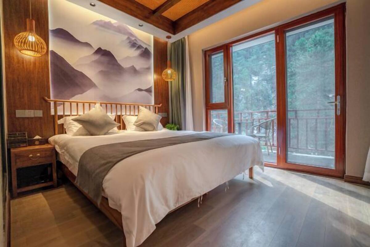 A bedroom with a bed, wooden headboard, nightstands, a wall mural, and a sliding glass door to a balcony.