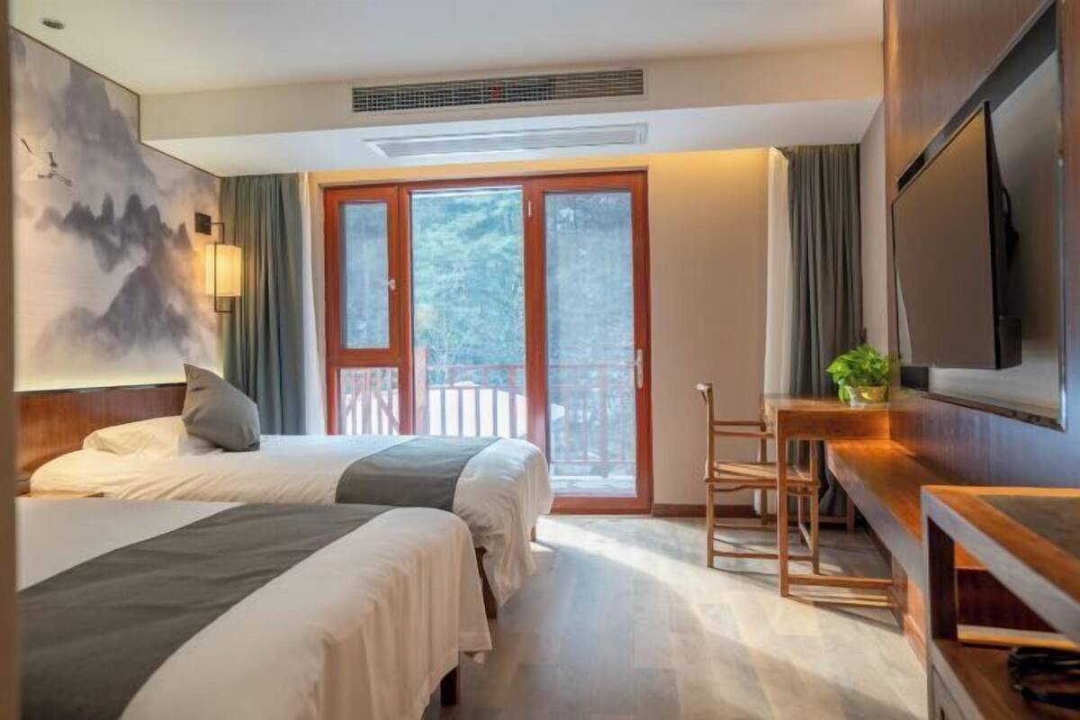 A hotel room with two beds, a desk, a TV, and a balcony with a view of trees.