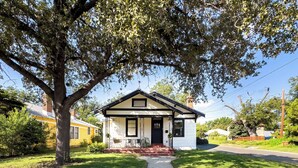 A single-story house with a black roof and white walls, a front porch with two chairs, and a large tree in front. - King William Charm 2-bedroom house in welcoming San Antonio with AC (San Antonio)