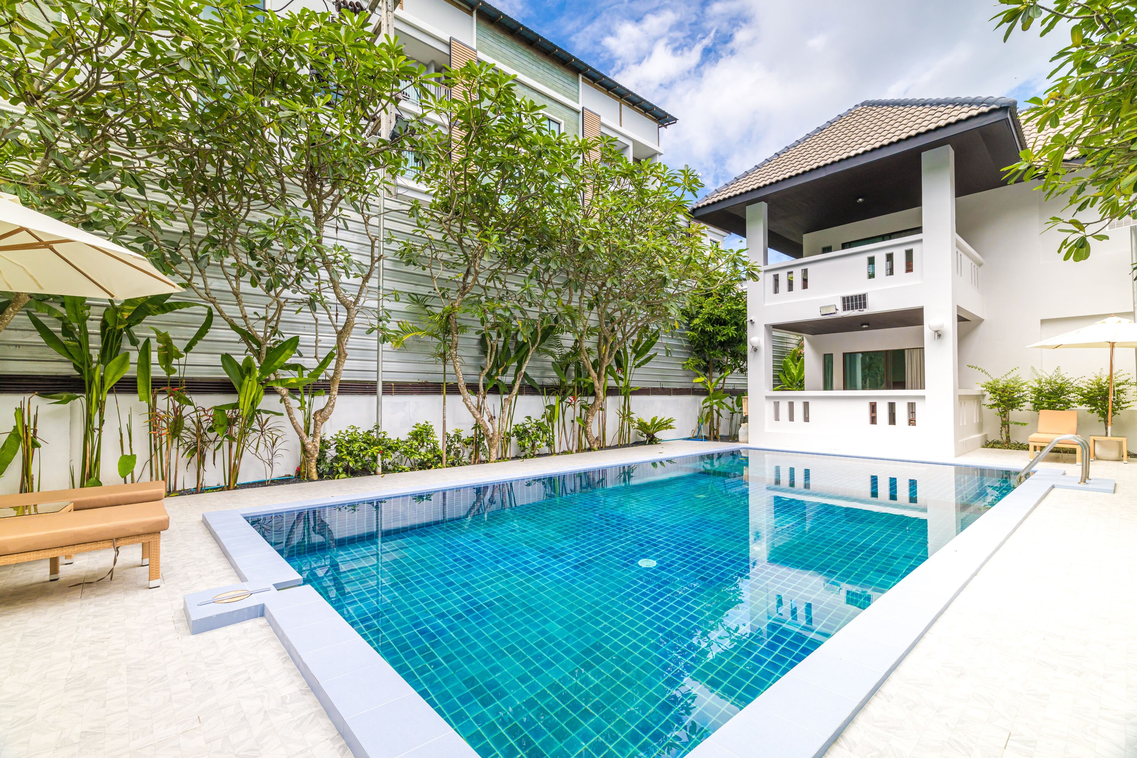 A swimming pool with tiled edges, surrounded by greenery and a white building with balconies.