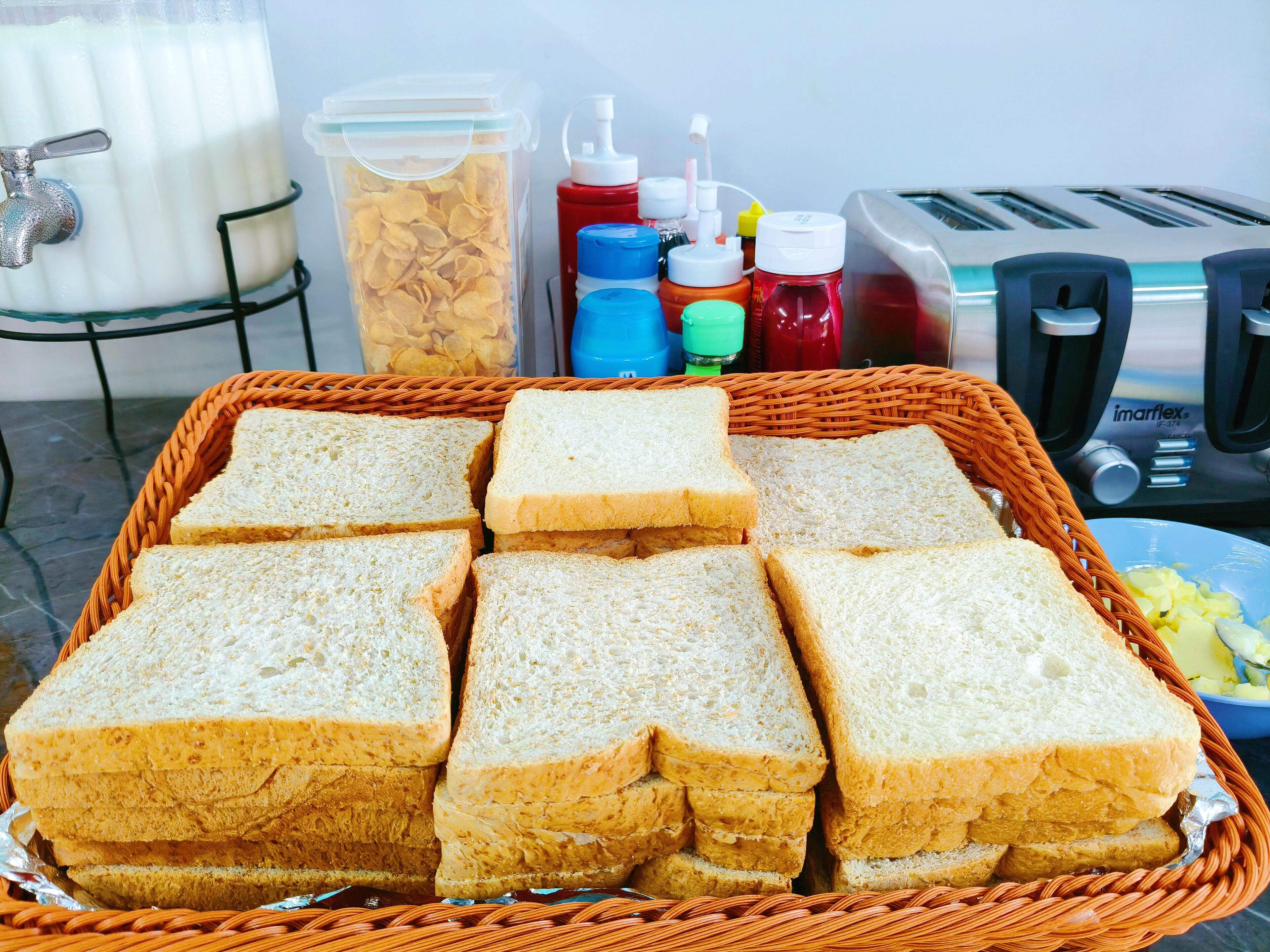 A basket of sliced bread, a milk dispenser, condiments, and a toaster on a table.