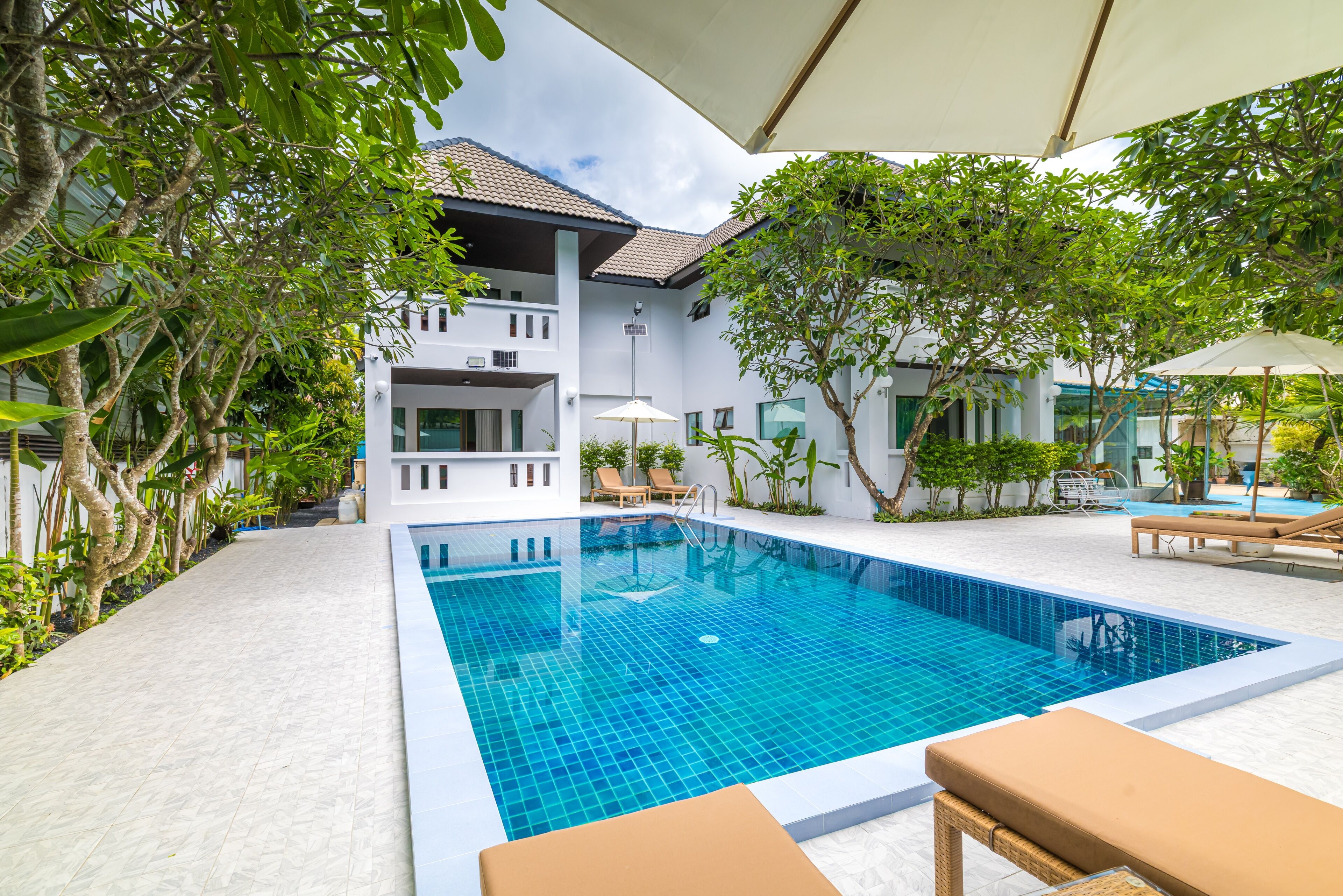 A swimming pool with a tiled deck, surrounded by lounge chairs and umbrellas, with a building in the background.