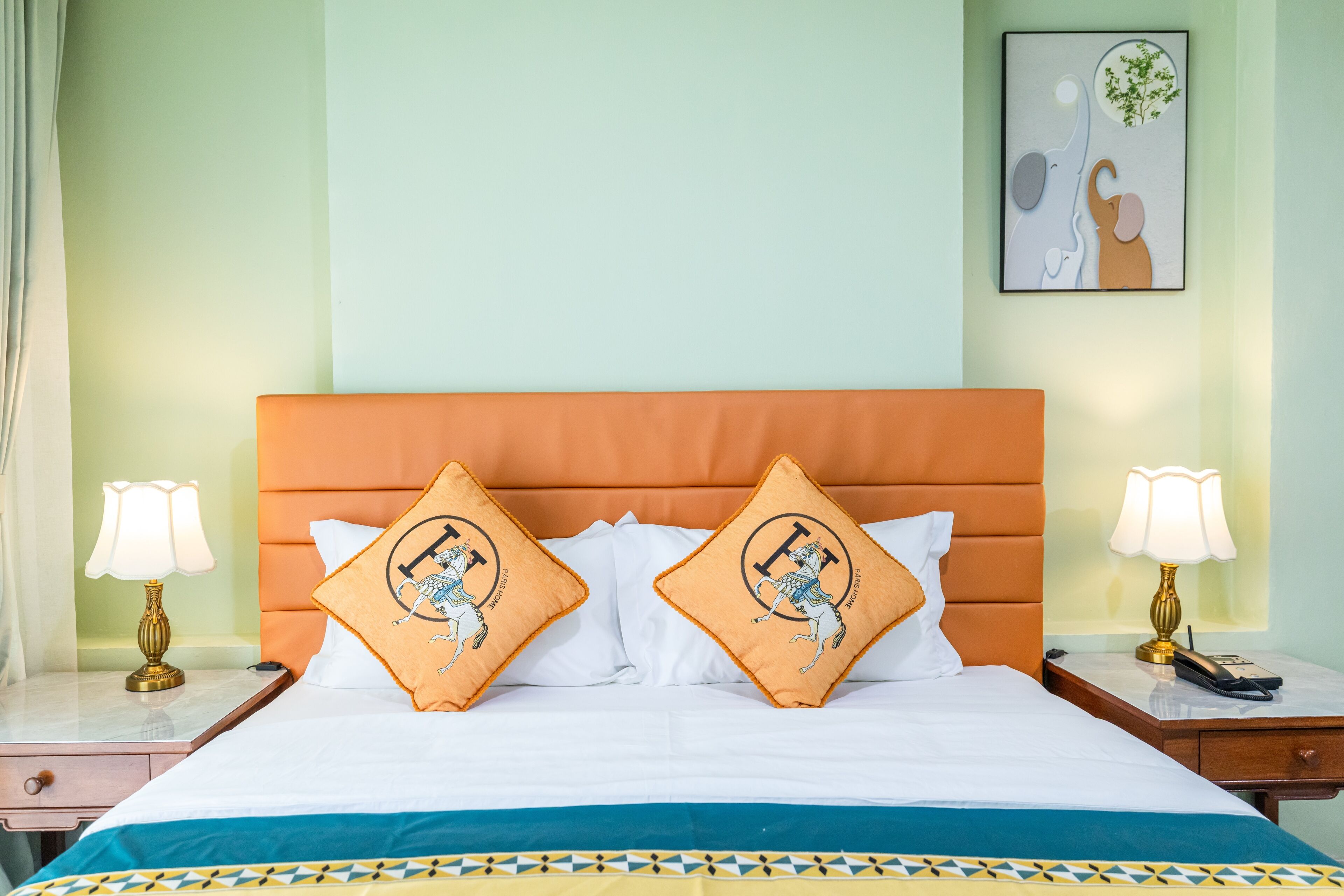 Una cama bien hecha con dos almohadones decorativos, una mesita de noche con una lámpara y una obra enmarcada en la pared.