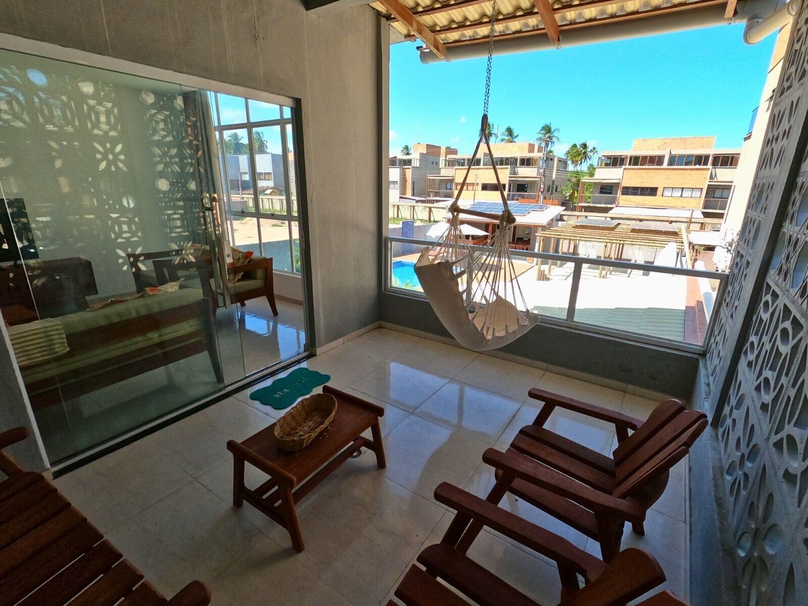 Une chambre moderne avec une porte en verre, un fauteuil suspendu, des meubles en bois et une vue sur une piscine et des bâtiments.