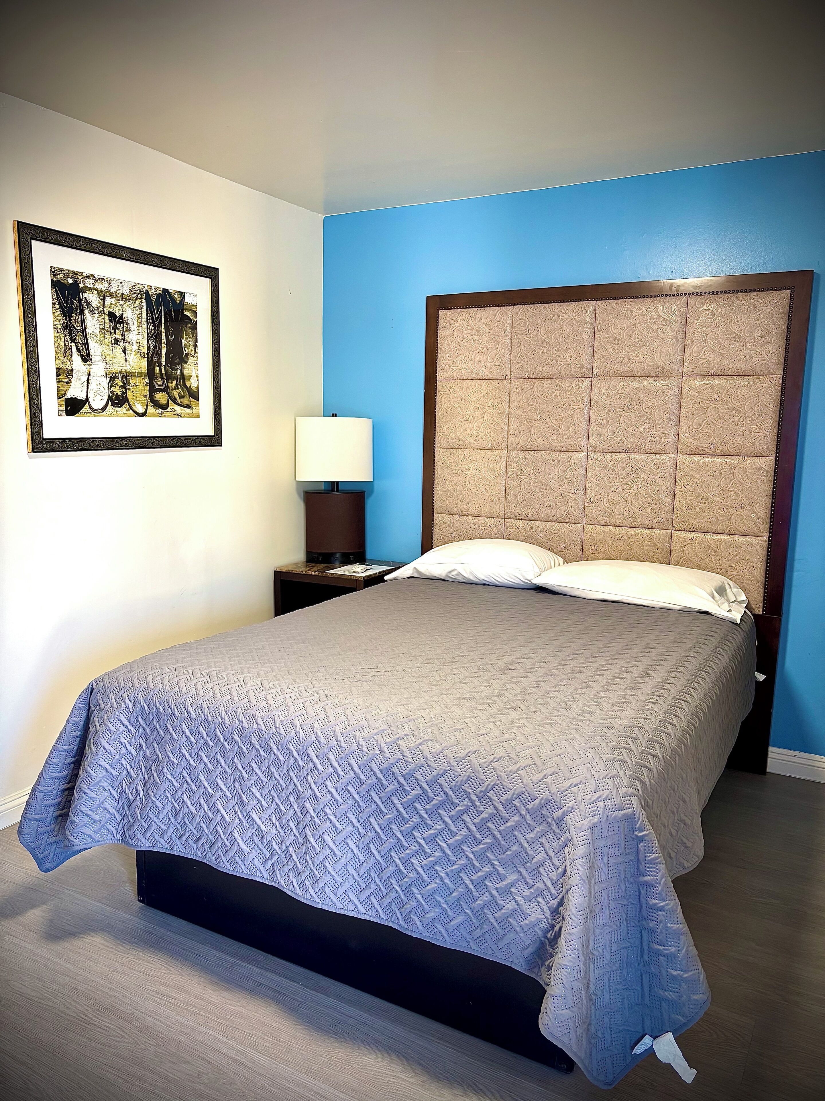 Een netjes opgemaakt bed met een beddekleed met een patroon, een hoofdbord met een structuur, een ingelijst kunstwerk aan de muur en een nachtkastje met een lamp.