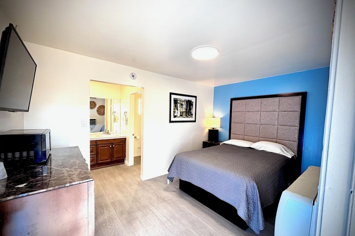 A bedroom with a bed, a dresser, a TV, and a bathroom visible in the background.