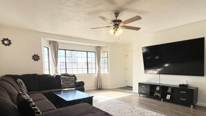 A living room with a sectional sofa, a coffee table, a flat-screen TV, a ceiling fan, and a large window with curtains. - Entire Newly Remodeled Home With Huge Parking (Quartz Hill)