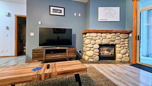 A living room with a stone fireplace, a flat-screen TV on a wooden stand, and a framed picture on the wall. - Rare Upper Village Townhouse w/a garage for gear and tuning! (Panorama)