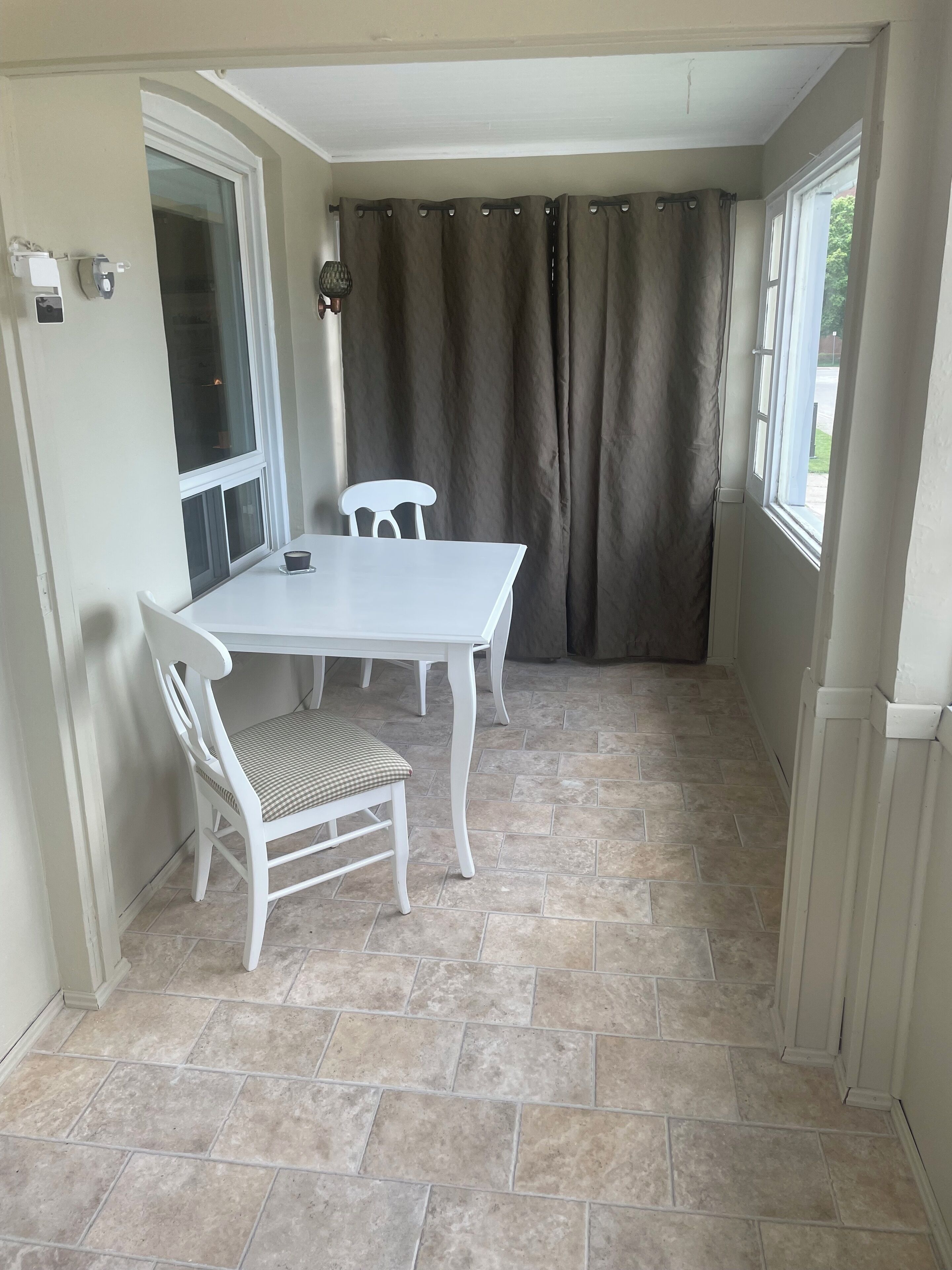 A small, enclosed patio area with a white table and two chairs, a window with a curtain, and a tiled floor.