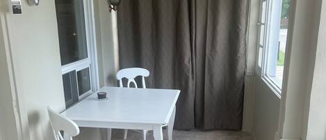 A small, enclosed patio area with a white table and two chairs, a window with a curtain, and a tiled floor.