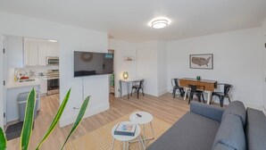 A modern living room with a grey sofa, a flat-screen TV, a dining area with a wooden table and chairs, and a kitchen with white cabinets and appliances. - Charming Modern Cottage, Downtown @pinpointstays (Coos Bay)