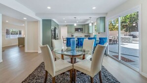 A modern kitchen with teal cabinets, a dining area with a glass table, and a view of a patio with outdoor furniture. - Hey! I Am Adriana On The Block | Pool | BBQ (Mission Viejo)