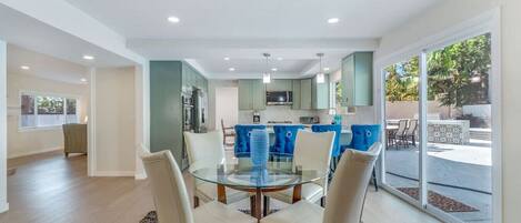Une cuisine moderne avec des meubles de couleur turquoise, un coin repas doté d’une table en verre et une vue sur un patio aménagé avec du mobilier d’extérieur.