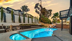 A swimming pool with a curved edge, surrounded by a concrete deck and a low wall with vertical plants. There are lounge chairs and a table on the deck, and a basketball hoop to the right. - Hey! I Am Adriana On The Block | Pool | BBQ (Mission Viejo)