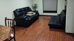 A living room with a black leather sofa, a wooden chair, a table with a lace tablecloth, a standing fan, and a window with a blind. - Charming 1-bedroom apartment in Surco with AC, two blocks from American Embassy (Santiago de Surco)
