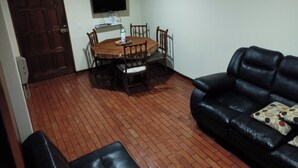 A living room with a dining table, chairs, a black leather sofa, a TV mounted on the wall, and a clock. - Charming 1-bedroom apartment in Surco with AC, two blocks from American Embassy (Santiago de Surco)