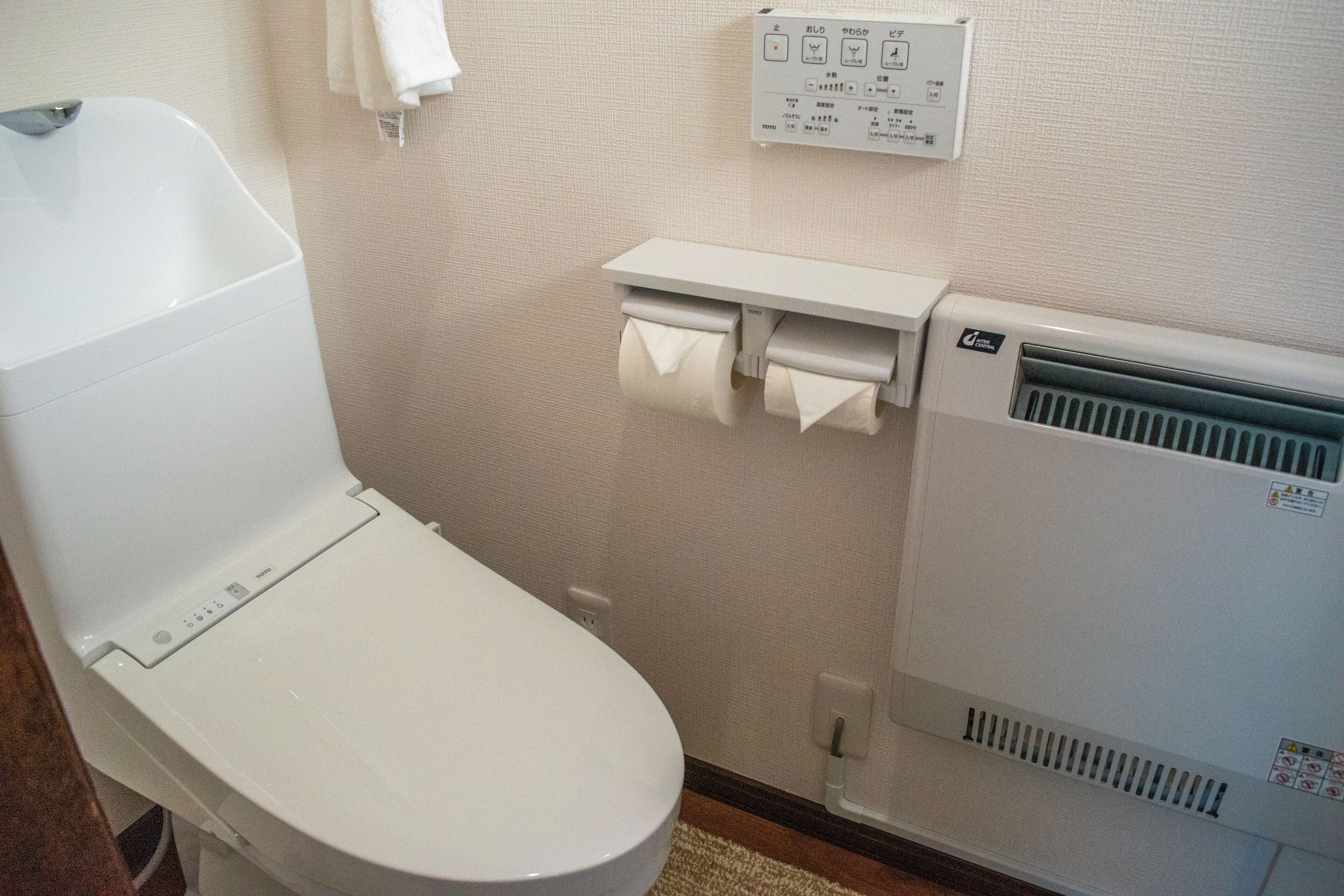A white toilet with a control panel, a paper towel dispenser, and a wall-mounted heater.