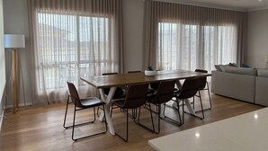 A modern living room with a wooden dining table, six chairs, a grey sofa, and large windows with sheer curtains.